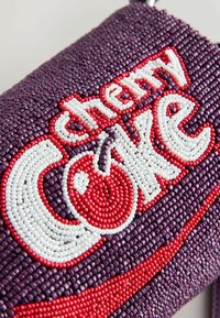 Gros plan sur une surface perlée de violet avec le texte "cherry coke" et un design de cerise rouge en perles blanches, rouges et violettes.