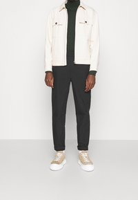 Crème kleurige jacket met twee borstzakken, donkergroene coltrui, zwarte broek en tan sneakers met witte zolen. Casual outfit.