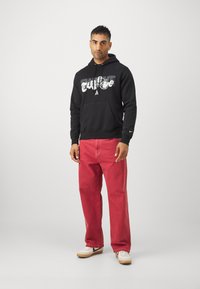 Felpa nera con cappuccio e testo grafico, abbinata a jeans rossi larghi e sneakers bianche con dettagli neri. Design semplice e stile casual.