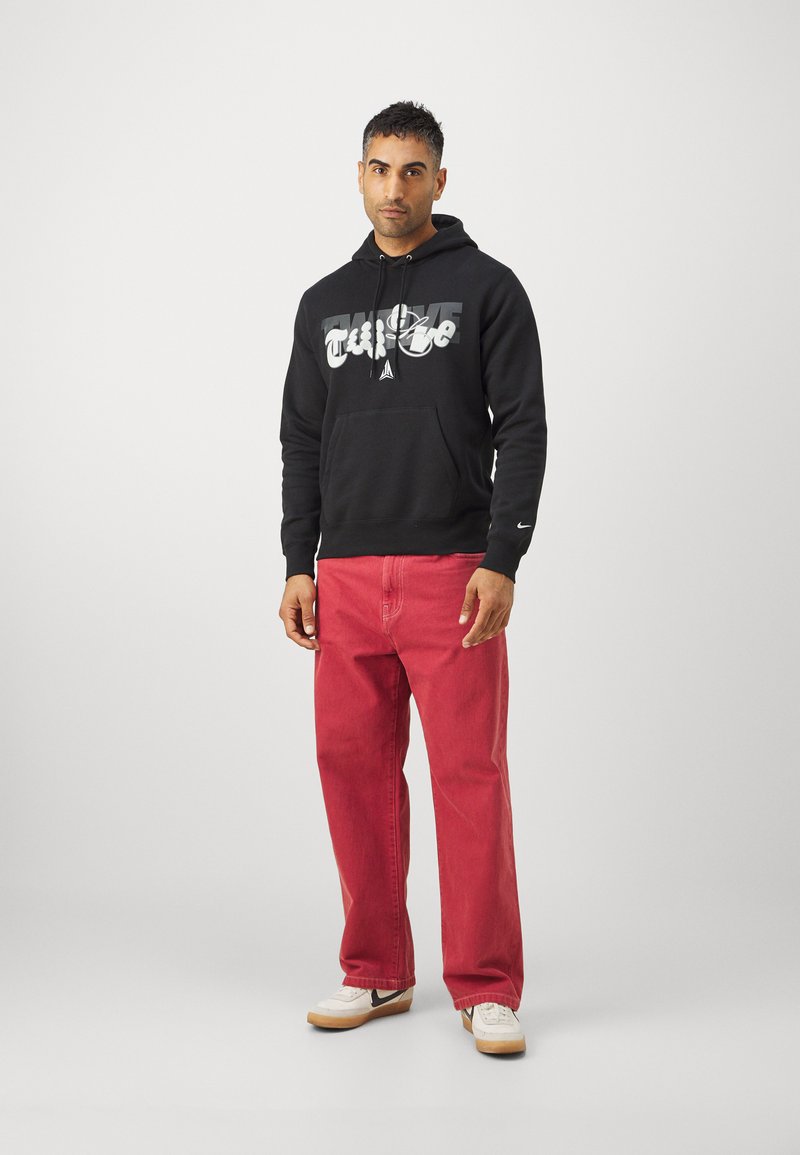 Czarna bluza z kapturem z graficznym napisem, w połączeniu z luźnymi czerwonymi dżinsami i białymi trampkami z czarnymi akcentami. Prosty design i casualowy styl.