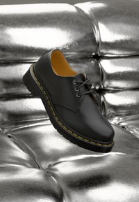 Černá kožená bota se žlutým prošíváním, designem na šněrování, kulatou špičkou a strukturovanou podrážkou, umístěná na lesklém, metalickém povrchu.