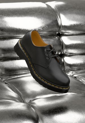 Zapato de piel negra con costuras amarillas, diseño con cordones, punta redondeada y suela texturizada, posicionado sobre una superficie metálica brillante.