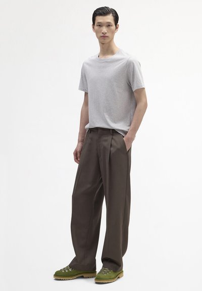 Jeune homme portant un t-shirt gris clair, un pantalon marron à jambes larges et des chaussures de randonnée vertes, debout avec une main dans la poche.