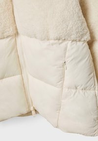 Jakke i cremefarve med en blød, struktureret overdel og en glat, quiltet nederste del med lynlås i siden til lukning.