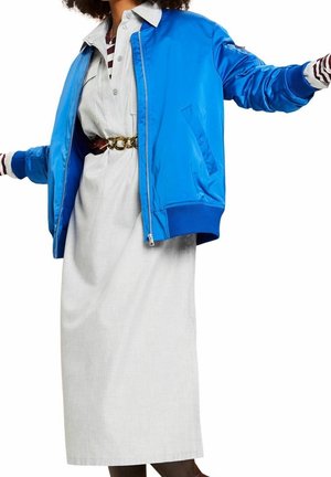 Blauwe satijnen bomberjack met geribbelde manchetten, gedragen over een lichtgrijze jurk met een gouden kettingriem. Een gestreept shirt steekt onder het jack uit.