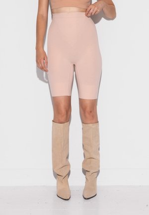 Vrouw die beige shorts met hoge taille en knielengte draagt en slonzige beige suède laarzen, staand tegen een witte muur.