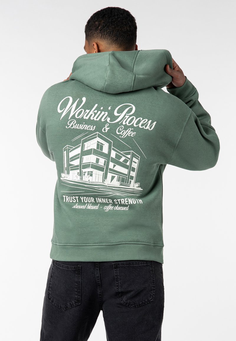 Grüne Kapuzenjacke mit einem großen weißen grafischen Motiv auf dem Rücken, das eine Gebäudeillustration und Text zeigt. Weicher Stoff, Kapuze mit Kordelzug und gerippte Bündchen.