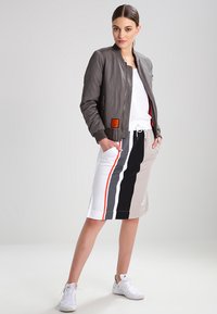 Graue Bomberjacke mit gerippten Bündchen, getragen über einem weißen, strukturierten Oberteil. Kombiniert mit einem knielangen Rock, der vertikale Streifen in Schwarz, Grau, Orange und Weiß aufweist.