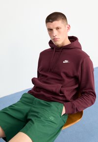 Sweatshirt bordô com bolso frontal e logotipo branco, combinado com calções verdes com um pequeno logotipo. Tecido suave, corte casual.