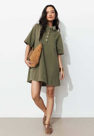 Femme portant une robe boutonnée à manches courtes vert olive, tenant un sac en daim beige, accessoirisée avec des bracelets dorés et des ballerines imprimé léopard.