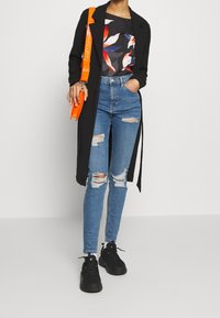 Abrigo negro, blusa floral, vaqueros desgarrados azul claro y zapatillas negras. La correa de la bolsa naranja contrastante añade un toque brillante.