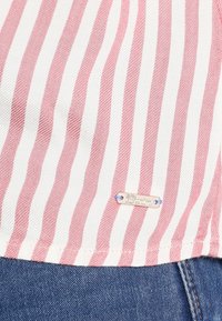 Koszula w paski z naprzemiennymi różowymi i białymi paskami, z małą metalową metką oznaczoną "Denim" u dołu. Teksturowany materiał.