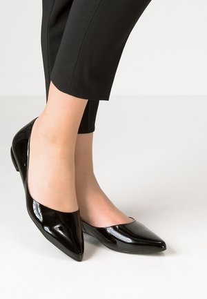 Pieds portant des ballerines noires en cuir verni à bout pointu, associées à un pantalon noir court, sur un fond clair.