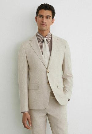 Fiatal férfi bézs kockás öltönyben, barna-fehér csíkos inggel és krémszínű nyakkendővel, fehér fal előtt áll.