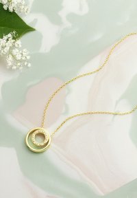 Guld vedhæng halskæde med to sammenflettede cirkulære former, hvoraf den ene er prydet med klare krystaller, på en fin guld kæde. Baggrunden har et marmor mønster.