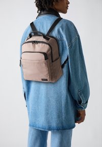 Zaino in nylon beige con due tasche frontali con zip e dettagli neri. Indossato sopra una giacca di jeans blu, presenta spallacci regolabili e una maniglia superiore.