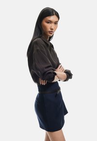 Chemise oversize marron foncé avec des manches retroussées, associée à une jupe navy taille haute avec une ceinture noire, sur un fond neutre.