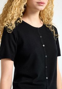 Černý krátký rukáv cardigan vyrobený z měkké pleteniny, s kulatým výstřihem a pěti malými černými knoflíky vpředu.