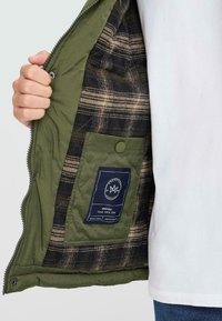 Giacca trapuntata verde oliva con fodera interna a quadri nera e beige, dotata di tasca con etichetta e chiusura frontale con zipper.