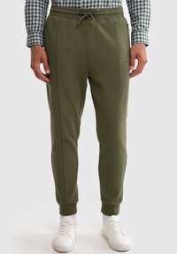 Olivegrønne joggere med snoretræk i taljen, sidesømme og ribkanter ved anklerne, lavet af blødt stof. Parret med hvide sneakers.