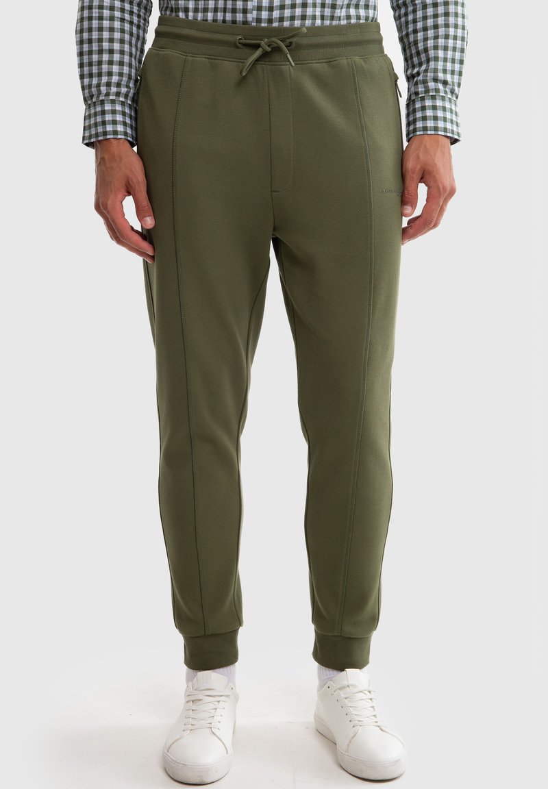 Olivegrønne joggere med snoretræk i taljen, sidesømme og ribkanter ved anklerne, lavet af blødt stof. Parret med hvide sneakers.