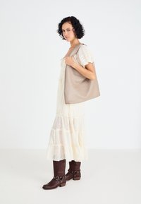 Beige strukturierte Tragetasche mit einem geometrischen Muster, über der Schulter getragen. Das Modell trägt ein cremefarbenes, gestuftes Kleid und braune Ankle Boots.
