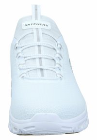 Λευκό αθλητικό παπούτσι με επάνω μέρος από δίχτυ, ελαστικά κορδόνια και μαξιλαρωμένη φτέρνα. Διαθέτει μια προφανή λογότυπο και σχέδιο επιφάνειας με υφή.