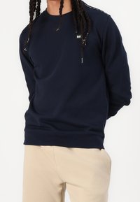 Personne aux longues tresses portant un sweat-shirt bleu marine et un pantalon beige clair, debout devant un fond blanc.