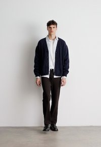 Sötétkék kardigán gombokkal, fehér és kék csíkos ing fölött viselve. Slim fit barna nadrág és fekete cipő teszi teljessé a megjelenést.