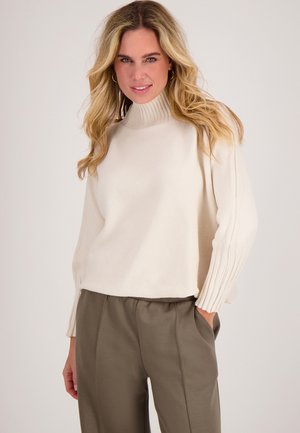 Crème-kleurige gebreide trui met een hoge ribbelkraag en ribbeldetails op de mouwen, gecombineerd met olijfgroene broek. Zachte textuur en een comfortabele pasvorm.