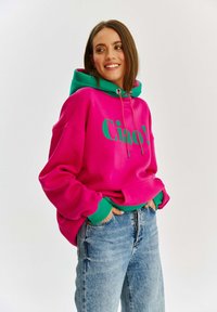 Jasnoróżowa bluza z kapturem z zielonymi akcentami i kieszenią z przodu, z napisem "Ciao!" w pogrubionej zielonej czcionce. Miękka tkanina i luźny krój.