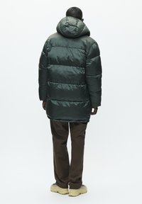 Un homme se tenant de dos, portant une veste matelassée vert foncé avec capuche, un pantalon marron et des chaussures de randonnée beiges sur fond blanc.