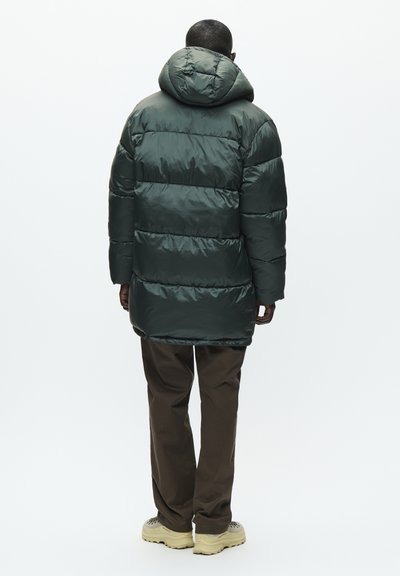 Un homme se tenant de dos, portant une veste matelassée vert foncé avec capuche, un pantalon marron et des chaussures de randonnée beiges sur fond blanc.