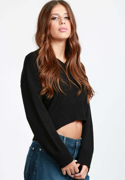 Giovane donna con lunghi capelli castani mossi, che indossa un maglione nero corto e jeans blu a vita alta, su uno sfondo semplice.