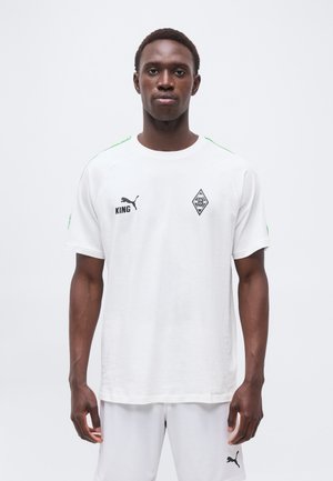 Άνδρας που φοράει λευκή αθλητική ένδυση με το λογότυπο Puma, το κείμενο "KING" και ένα διαμαντόσχημο έμβλημα σε μονόχρωμο φόντο, κοιτάζοντας μπροστά με ουδέτερη έκφραση.