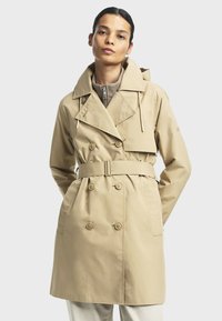 Žena v béžovom dvojradovom trench coat s opaskom, oblečená do svetlého zipsového topu, stojí na svetlom pozadí.