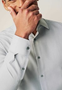 Hellgraues Hemd aus texturiertem Stoff, mit Ein-Knopf-Manschetten und dunkelgrünen Knöpfen. Das Modell fasst sich ans Gesicht und zeigt den Kragen des Hemdes.