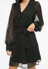 Czarna sukienka w białe groszki z kołnierzykiem, zapinana na guziki z przodu, z długimi przejrzystymi rękawami. Posiada pasek wiązany w talii oraz rozkloszowaną spódnicę.