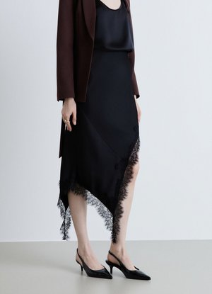 Donna con gonna nera asimmetrica rifinita in pizzo, top nero, blazer marrone scuro e scarpe slingback nere con tacco, in piedi su sfondo semplice.
