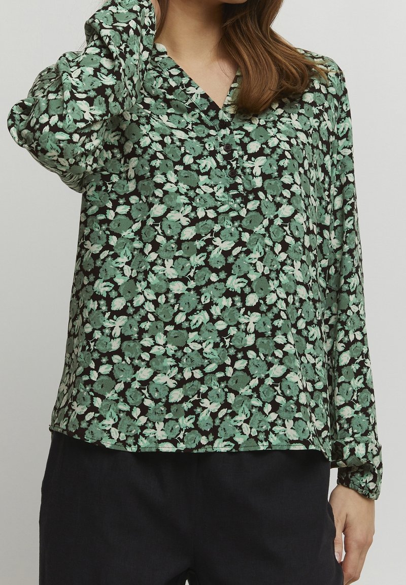 Blusa floreale con sfondo nero, decorata con motivi floreali verdi e bianchi, maniche lunghe e scollatura a V con bottoni.