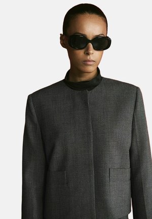 Woman with slicked-back hair wearing oversized black sunglasses and a gray textured blazer with a high neckline.