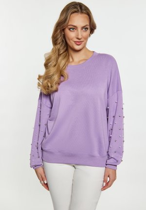 Kvinde med langt blondt hår iført en lavendelfarvet sweater med perle- og palietdetaljer på ærmerne og hvide bukser, der står mod en ensfarvet baggrund.