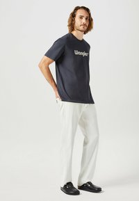 Mörkblå t-shirt med "Wrangler"-logotyp, kombinerad med ljusfärgade byxor och svarta träskor. Mjuk tygkvalitet, avslappnad passform, korta ärmar.