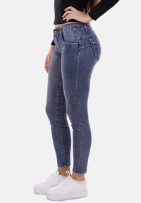 Jeans skinny blu in denim, con vita alta media, due tasche posteriori e una leggera texture sbiadita. Abbinati a scarpe da ginnastica bianche.