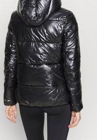 Svart pufferjacka med glänsande, vadderad utsida, som har huva och figurnära silhuett. Texturerat, lättviktsmaterial med horisontella sömmar.