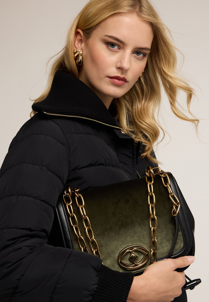Giacca imbottita nera con colletto alto, che tiene una borsa in suede verde con tracolla a catena dorata e un dettaglio dorato sul davanti.