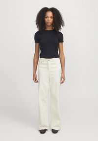 Top preto de mangas curtas combinado com calças largas creme; as calças têm uma cintura alta, costuras visíveis e fecho com botão. Sapatos pretos completam o visual.