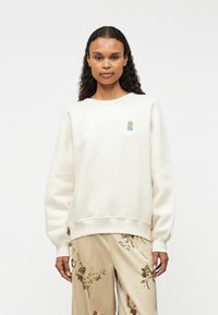 Krém színű pamut pulóver bordázott mandzsettákkal és aljjal, a mellrészen egy kis, színes logóval. Puha tapintású és laza állású.