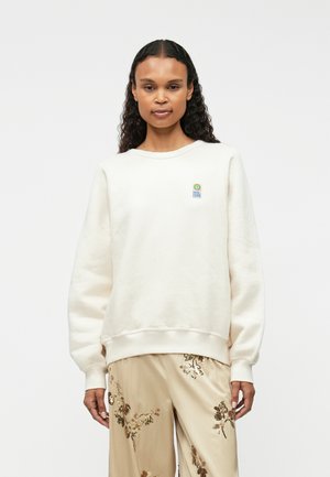 Krém színű pamut pulóver bordázott mandzsettákkal és aljjal, a mellrészen egy kis, színes logóval. Puha tapintású és laza állású.