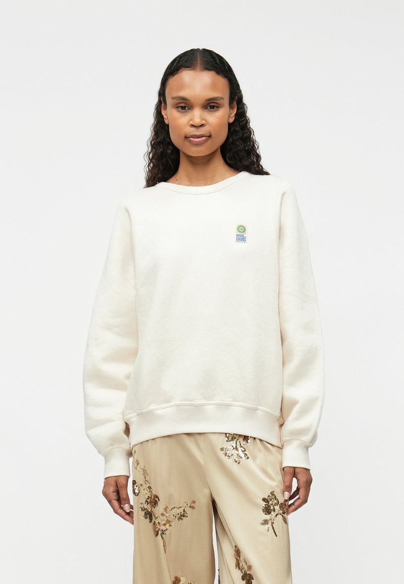 Krém színű pamut pulóver bordázott mandzsettákkal és aljjal, a mellrészen egy kis, színes logóval. Puha tapintású és laza állású.